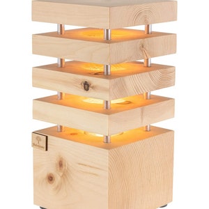 Könnte beinhalten: Eine Holztischlampe mit quadratischem Sockel und vier übereinander gestapelten Schichten. Die Lampe hat ein warmes, gelbes Licht, das durch das Holz scheint. Die Lampe ist aus Naturholz gefertigt und hat ein schlichtes, modernes Design. Die Lampe ist mit "Holz Glanz" beschriftet.