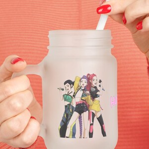 Peut inclure: Bocal en verre dépoli avec une poignée, orné d'un graphisme coloré de trois personnages avec des microphones. Une paille blanche dépasse du bocal. Le bocal est tenu par une personne avec du vernis à ongles rouge.