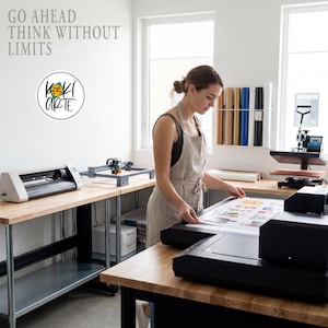 K&ouml;nnte beinhalten: Eine Frau in einer Werkstatt, die eine Sch&uuml;rze tr&auml;gt, untersucht einen Druck. Der Arbeitsbereich umfasst eine Hei&szlig;presse, eine Schneidemaschine und Vinylrollen. Der Text an der Wand lautet "GO AHEAD THINK WITHOUT LIMITS" und ein Logo "KOKI ARTE".