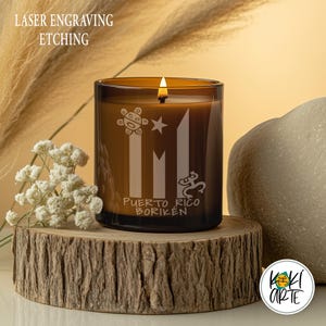 K&ouml;nnte beinhalten: Eine brennende braune Glas-Kerze mit lasergravierten puerto-ricanischen Symbolen und dem Text "Puerto Rico Borik&eacute;n". Die Kerze steht auf einem Holzsockel mit wei&szlig;en Blumen und einem Stein im Hintergrund. Der Text oben lautet "LASER ENGRAVING ETCHING".