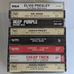 Puede incluir: Una pila de cintas de casete vintage con varios artistas y álbumes, incluyendo Elvis Presley, Deep Purple y Judas Priest. Las cintas tienen etiquetas con nombres de artistas, títulos de álbumes y números de catálogo. Las cintas están en estuches de plástico transparente.