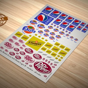 Puede incluir: Una hoja de papel de calcomanía blanco con varios logotipos de bebidas gaseosas impresos en ella. Los logotipos incluyen Pepsi, Fanta, Schweppes y Dr Pepper. El texto "Matho Models 80021 Soft Drink Logos B - modern - all scales" está impreso en la parte inferior de la hoja.