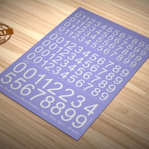 May include: A sheet of white number decals on a lavender background. The numbers 0-9 are repeated in multiple rows. The text "Matho Models 80004 Decal Numbers - large, white, type 1 - all scales" is printed at the bottom.