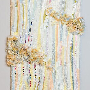 May include: A white tapestry with a vertical striped pattern made from strips of fabric in various shades of pink, yellow, blue, and white. The fabric is frayed and textured, creating a unique and rustic look.