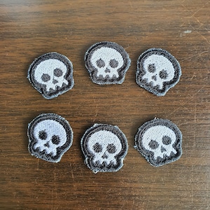 May include: Six small black and white skull patches with a white outline. The patches are on a brown wooden surface.
