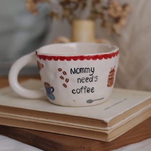 May include: White ceramic coffee mug with a red scalloped rim, featuring the text "Mommy needs coffee" and coffee bean illustrations. The mug is resting on a stack of books and a wooden board.