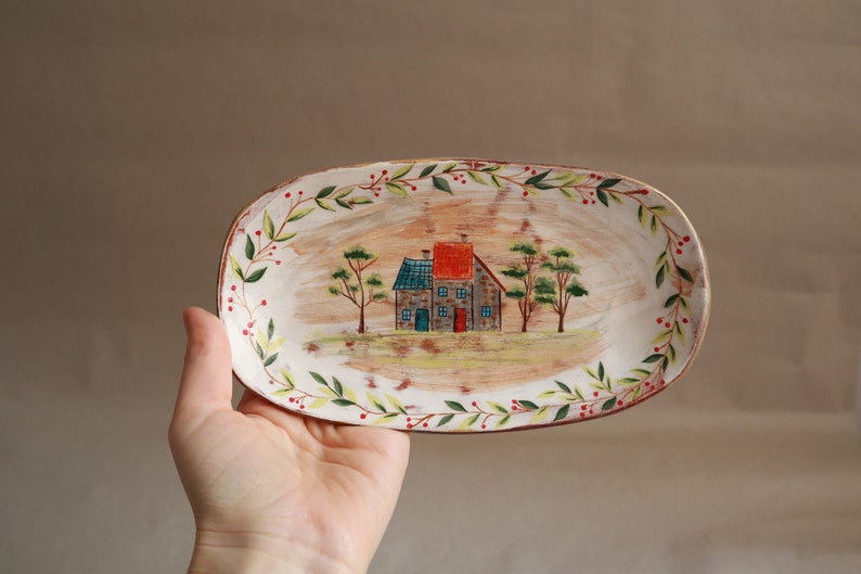 Pu&ograve; includere: Un piatto ovale in ceramica bianca con una scena dipinta di una piccola casa con un tetto rosso e alberi verdi. Il piatto &egrave; decorato con un bordo floreale verde e rosso.