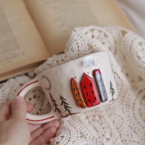 May include: Cream-colored ceramic mug with a whimsical village scene. The mug features three raised, colorful buildings, trees, and a crescent moon design. The handle has a red stripe. The mug is held over a white lace cloth.