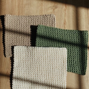 May include: Three crocheted washcloths in beige, green, and white are arranged on a wooden surface. The washcloths have a textured, woven appearance. The image is lit by natural light, casting shadows.
