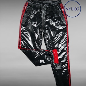 Puede incluir: Pantalones deportivos negros con rayas laterales y detalles en rojo. Los pantalones tienen un aspecto brillante y húmedo y cuentan con cremallera y bolsillos. La marca "NYLKO" es visible en un círculo azul.