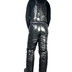 May include: Black, shiny snow bib overalls with adjustable straps and a quilted design. The overalls have a high-shine finish and are designed for cold weather activities. The bibs are shown from the back, with a black long-sleeve shirt underneath.