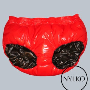 Op de afbeelding: Rode, glanzende, waterdichte onderbroek met zwarte panelen. De elastische tailleband en beenopeningen zijn zichtbaar. De merknaam "NYLKO" staat in een zwarte cirkel rechtsonder.