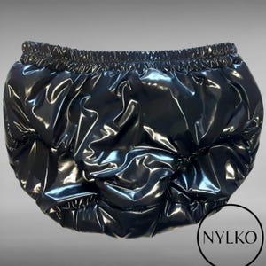 May include: A pair of black, high-gloss, vinyl-like bloomers featuring a gathered elastic waistband and leg openings. The fabric has a reflective, shiny finish with deep folds and creases throughout. A circular logo with the text NYLKO is visible.