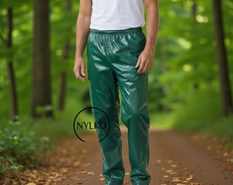 May include: Green waterproof pants with an elastic waistband. The pants are shiny and appear to be made of a synthetic material. The person is wearing a white t-shirt and white sneakers. The background is a blurred forest scene.