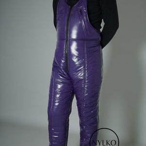 May include: Purple snow bib overalls with a shiny, quilted texture. The overalls have a full-length zipper, adjustable straps, and a small zippered pocket on the chest. The brand name "NYLKO" is visible on the lower right.
