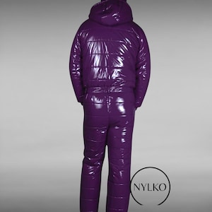 Puede incluir: Un mono acolchado de color morado intenso y alto brillo que incluye una chaqueta con capucha y pantalones a juego. El material tiene un acabado brillante y reflectante. Un logotipo circular con el texto NYLKO es visible abajo a la derecha.