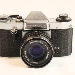 May include: A silver and black Praktica camera with a Pentacon Auto 1.8/50 lens. The camera has a black leatherette covering and a silver metal body. The lens has a black metal barrel and a silver metal ring.