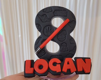 May include: Black and red Star Wars themed cake topper with the number 8 and the name Logan. The number 8 is decorated with Star Wars characters and symbols. The name Logan is in red letters with black outlines.
