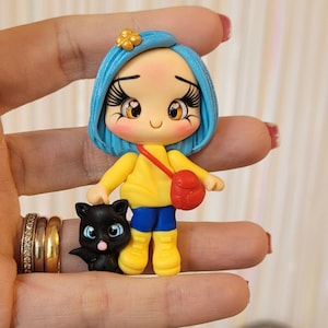 Könnte beinhalten: Handgefertigte Figur einer Figur mit blauen Haaren, einem gelben Oberteil, blauen Shorts und gelben Stiefeln, die eine rote Tasche trägt. Eine schwarze Katzenfigur befindet sich neben der Figur. Die Figur hat große Augen und eine Blume im Haar.