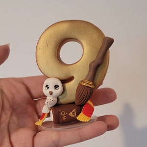 Op de afbeelding: Een gouden nummer negen taarttopper met een bruine en zwarte bezem, een witte uil en een rood en geel boek. De topper staat op een doorzichtige plastic basis.