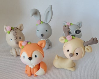 Op de afbeelding: Set van vijf bosdierenfiguren gemaakt van polymer clay. De figuren zijn geschilderd in tinten grijs, bruin en oranje. Elk figuur heeft een klein bloemetje op zijn hoofd. De dieren omvatten een konijn, een vos, een hert, een wasbeer en een eekhoorn.