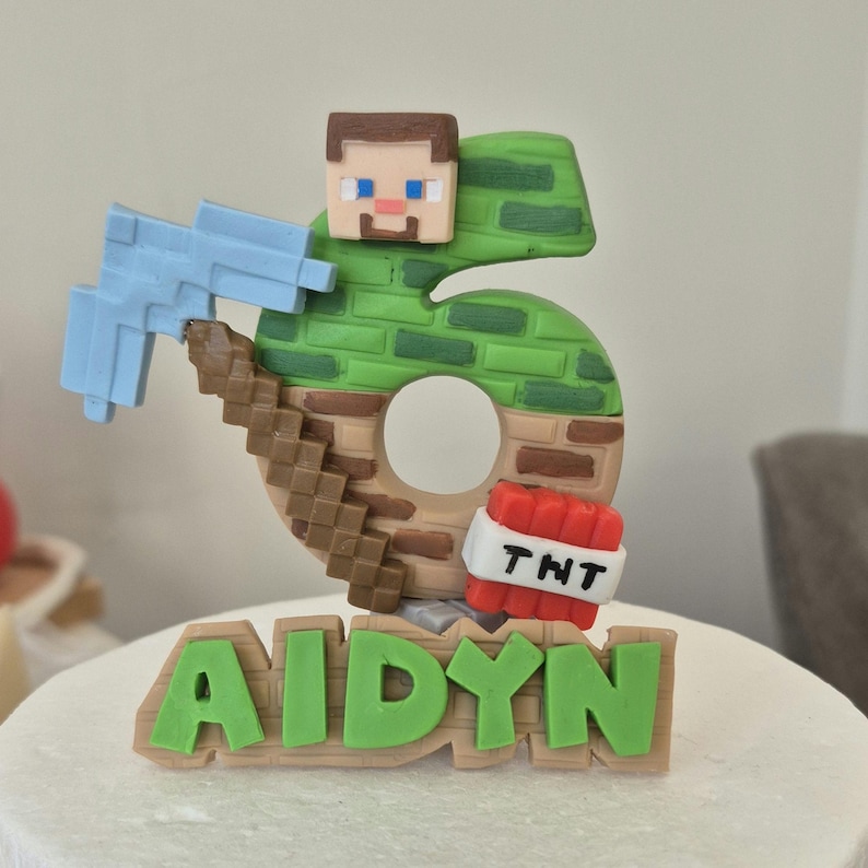 May include: A green and brown Minecraft-themed cake topper with a number 6, a figure of Steve, a pickaxe, and TNT. The name "AIDYN" is written in green letters on a brown base.