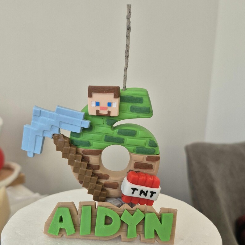 May include: A green and brown Minecraft-themed birthday cake topper with a number 6, a pickaxe, a TNT block, and the name "AIDYN" in green letters.
