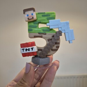 May include: A green and brown Minecraft-themed cake topper shaped like the number 5. The topper features a Minecraft character, a pickaxe, and a TNT block with the text "TNT" on it.