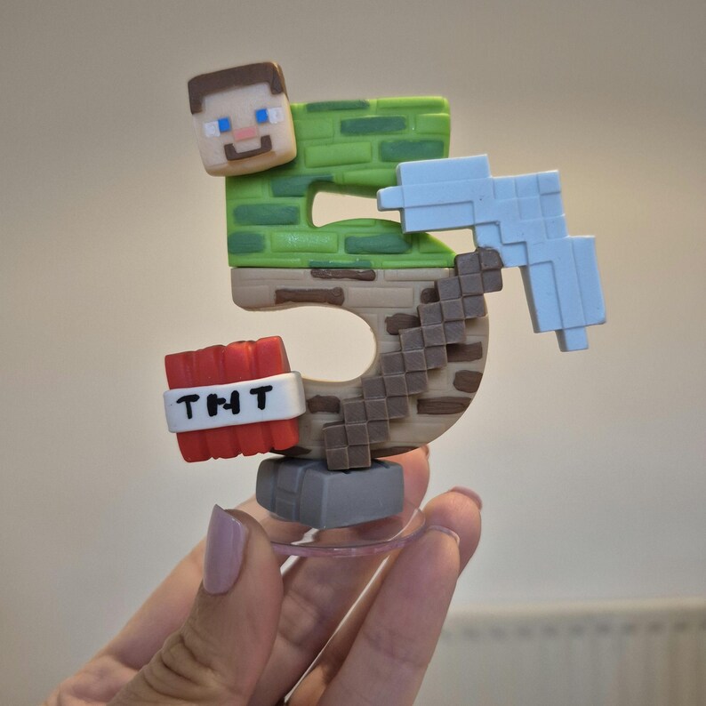 May include: A green and brown Minecraft-themed cake topper shaped like the number 5. The topper features a Minecraft character, a pickaxe, and a TNT block with the text "TNT" on it.