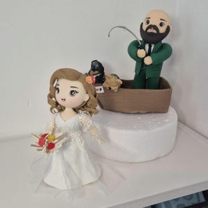 以下が含まれることがあります： 白いドレスを着てブーケを持ち、緑色のスーツを着た新郎がボートから釣りをするウェディングケーキトッパー。ボートには2つの小さな犬の置物が入っています。花嫁は茶色の巻き毛で、新郎はひげを生やしています。
