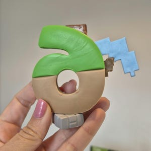 May include: A green and brown number six figurine with a blue and brown block attached to the side. The figurine is made of clay and has a textured surface.