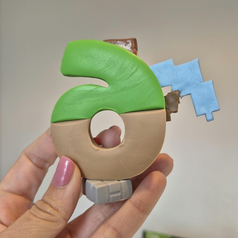 May include: A green and brown number six figurine with a blue and brown block attached to the side. The figurine is made of clay and has a textured surface.