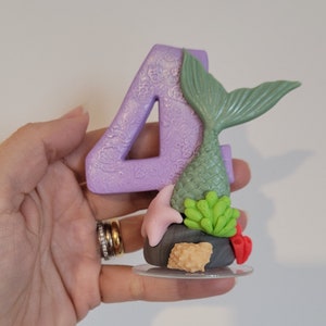 Puede incluir: Topper de pastel de número cuatro morado con una cola de sirena verde, conchas rosadas, algas verdes y una base de roca marrón.