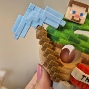 May include: A Minecraft character figurine holding a blue pickaxe. The character is wearing a green shirt and brown pants. The figurine is made of plastic and is about 6 inches tall.