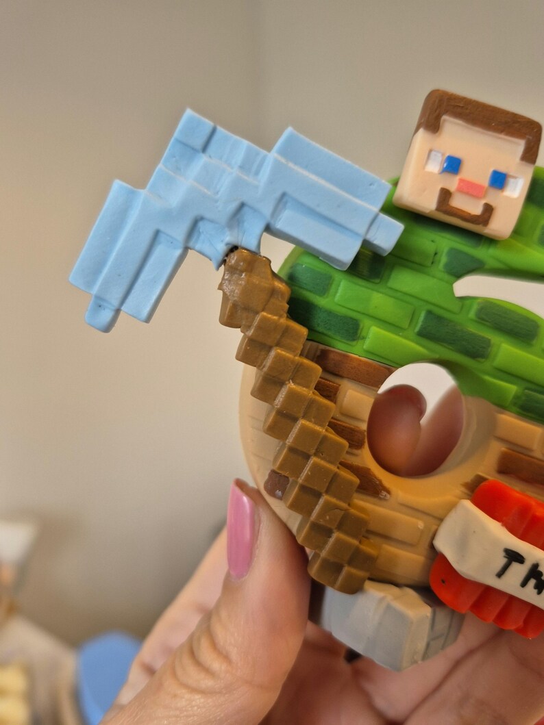 May include: A Minecraft character figurine holding a blue pickaxe. The character is wearing a green shirt and brown pants. The figurine is made of plastic and is about 6 inches tall.