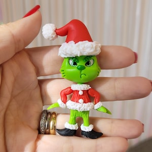Puede incluir: Un personaje verde de Grinch con un gorro de Papá Noel rojo y blanco y un suéter navideño rojo y blanco. El Grinch está hecho de arcilla polimérica y tiene una expresión gruñona.