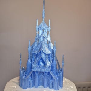 May include: A light blue, 3D-printed castle model with intricate architectural details. The castle is perched atop a white, snow-like base. The design features multiple turrets, spires, and detailed facades, creating a fantasy theme.