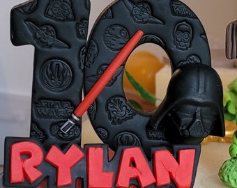 May include: Black and red Star Wars themed cake topper with the number 10 and the name Rylan. The number 10 is decorated with images of Yoda, Darth Vader, and the Death Star.