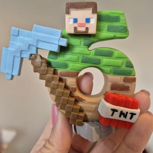 May include: A green and brown Minecraft-themed cake topper shaped like the number 6. The topper features a Minecraft character holding a pickaxe and a TNT block.