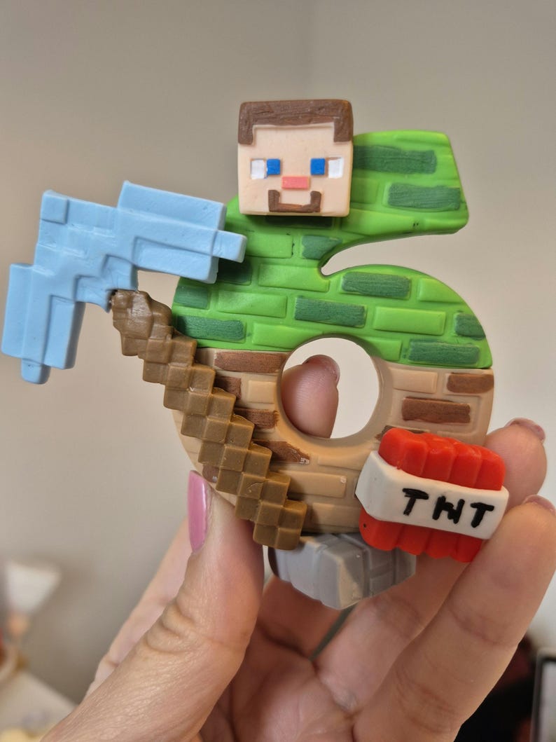 May include: A green and brown Minecraft-themed cake topper shaped like the number 6. The topper features a Minecraft character holding a pickaxe and a TNT block.