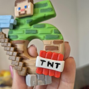 May include: A green and brown Minecraft-themed number 5 cake topper with a small figure of Steve holding a pickaxe. The number 5 is made to look like a block of dirt with a TNT block attached to the side. The TNT block has a white label with the letters "TNT" written in black.