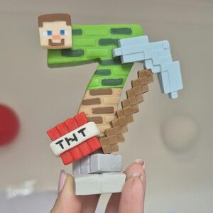 May include: A green and brown Minecraft-themed cake topper shaped like the number seven. The topper features a pixelated Minecraft character, a pickaxe, and a TNT block with the text "THT".