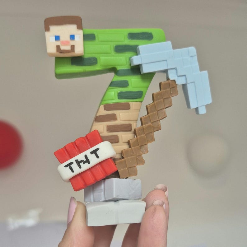 May include: A green and brown Minecraft-themed cake topper shaped like the number seven. The topper features a pixelated Minecraft character, a pickaxe, and a TNT block with the text "THT".