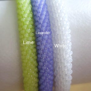 May include: Close-up of three beaded bracelets in lime green, lavender, and white. The bracelets are labeled with their respective colors. The beads have a textured, rounded appearance. The bracelets are displayed against a soft, blurred background.
