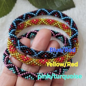 Puede incluir: Cuatro pulseras de cuentas en combinaciones de colores azul/rojo, amarillo/rojo y rosa/turquesa. Las pulseras están hechas de pequeñas cuentas de colores, y el texto de la imagen indica las combinaciones de colores. Las pulseras se muestran en una mano.