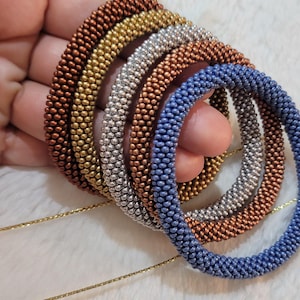 Könnte beinhalten: Eine Sammlung von Armbändern aus Perlen in verschiedenen Farben, darunter Bronze, Gold, Silber und Blau. Die Armbänder bestehen aus kleinen, runden Perlen und werden auf einer weichen, weißen Oberfläche präsentiert. Eine goldene Kette ist sichtbar.