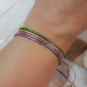 May include: A stack of beaded bracelets in shades of green, gold, pink, and silver. The bracelets are strung on thin cords and are worn on the wrist. The bracelets are a simple, elegant accessory.