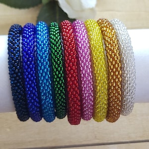 May include: A collection of beaded bracelets in various colors, including navy, blue, green, red, pink, yellow, and silver. The bracelets are displayed on a white cylinder against a wooden background, with a white rose in the background.