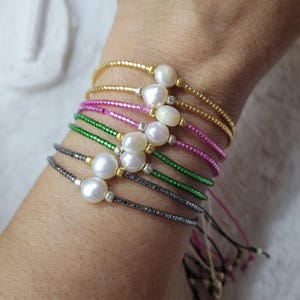 Può includere: Una collezione di braccialetti di perline in oro, rosa, verde e grigio scuro. Ogni braccialetto presenta una grande perla bianca. I braccialetti sono esposti su un polso, mostrando la varietà di colori e design.