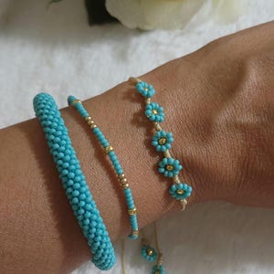 May include: Three beaded bracelets in shades of turquoise and gold. One bracelet is a thick, solid turquoise cylinder. Another is a thin strand of turquoise beads with gold accents. The third features turquoise flower-shaped beads.